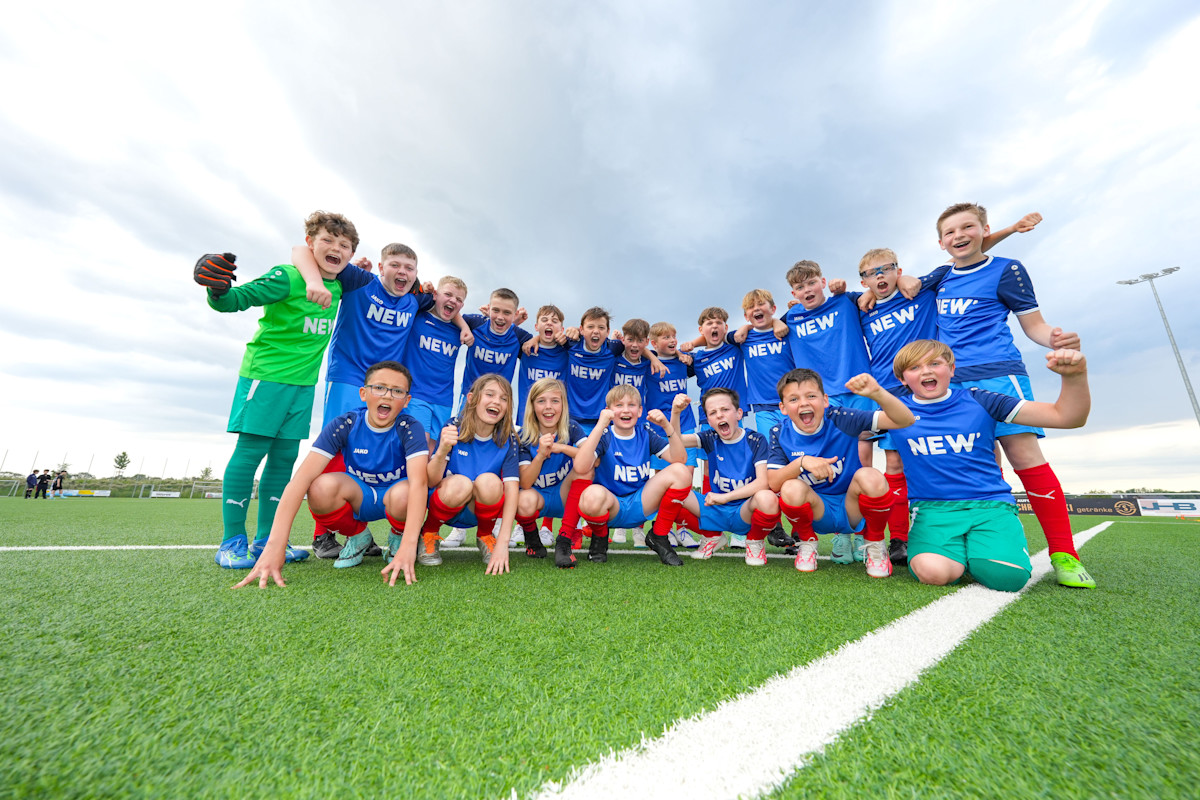 Eine Fußballmannschaft in blauen Trikots und roten Hosen posiert gemeinsam für ein Gruppenfoto auf einem grünen Fußballplatz. Die Spieler sind in zwei Reihen aufgestellt, wobei einige knien und andere stehen, unter einem bewölkten Himmel.