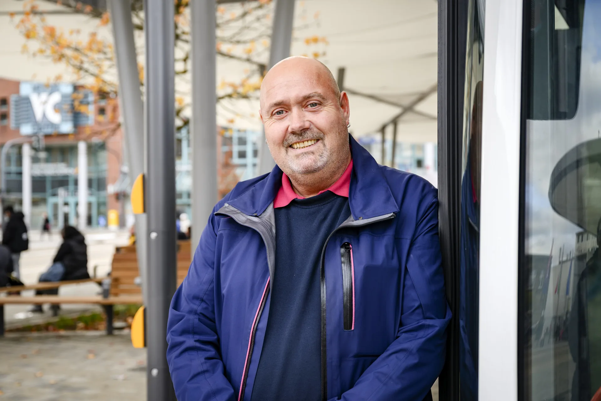 Ein freundlich lächelnder Mann in blauer Arbeitsjacke mit roten Akzenten steht an der Tür eines Fahrzeugs an einer Haltestelle. Im Hintergrund sind herbstliche Bäume mit orangefarbenem Laub, moderne Gebäude und wartende Fahrgäste zu sehen.