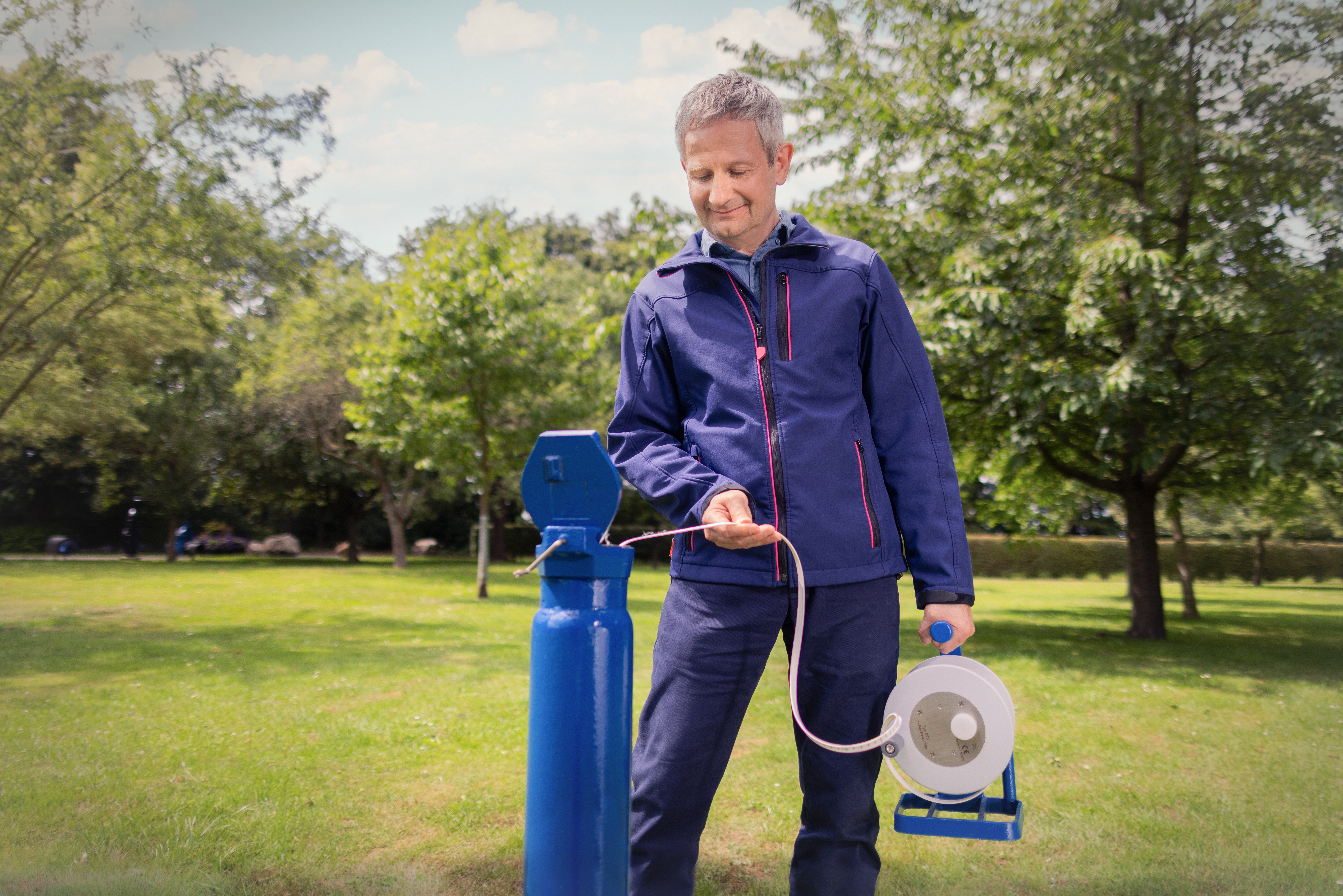 Eine Person in blauer Arbeitskleidung entnimmt Wasser an einer öffentlichen Trinkwasserstation. In der Hand hält sie eine Kabeltrommel, im Hintergrund sind eine Grünfläche und Bäume zu sehen.