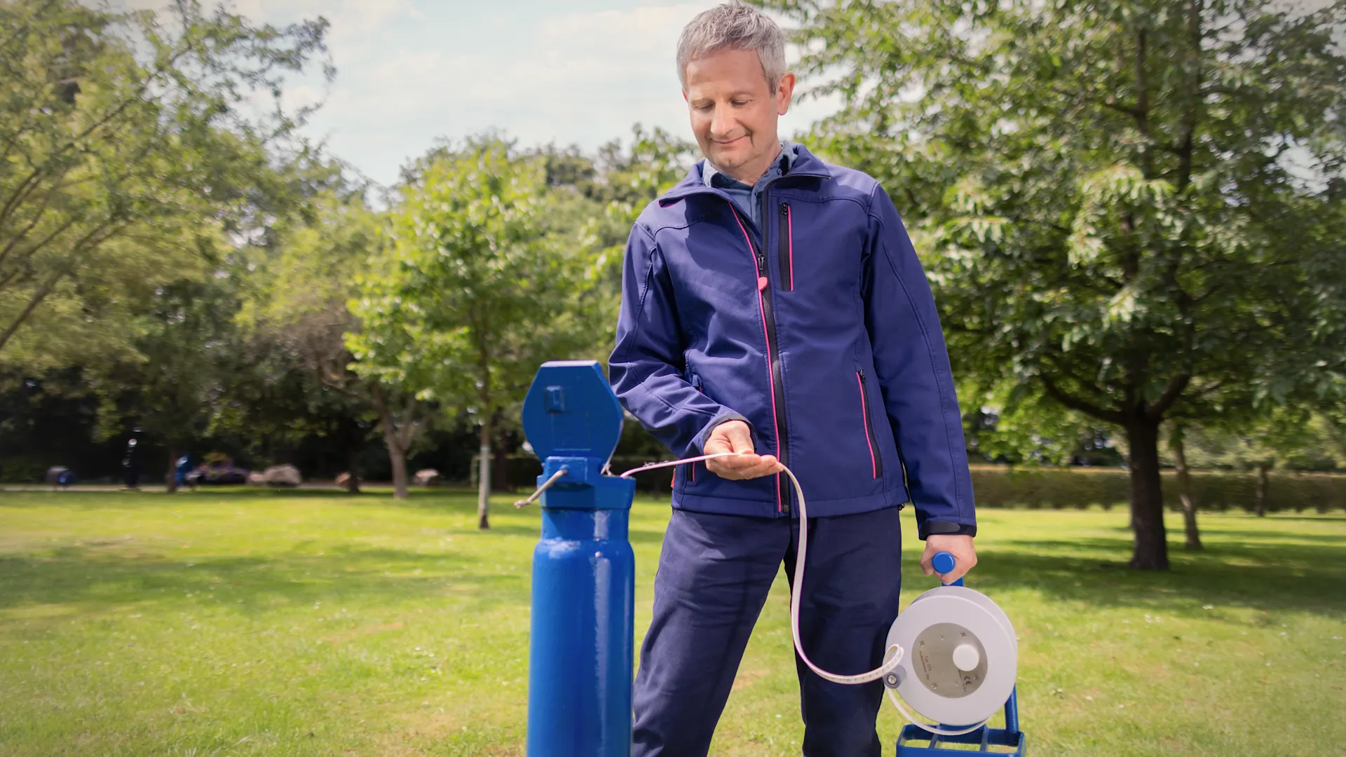 Ressourcen bewahren, Wasser schützen. Eine Person in blauer Arbeitskleidung entnimmt Wasser an einer öffentlichen Trinkwasserstation. In der Hand hält sie eine Kabeltrommel, im Hintergrund sind eine Grünfläche und Bäume zu sehen.