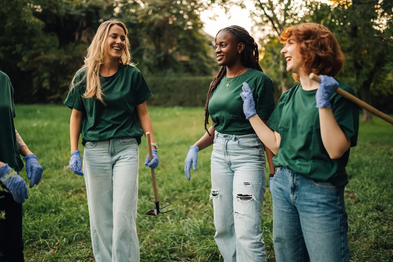 Freiwilligendienst in Europa: Reisen mit Sinn und Engagement Das Bild zeigt drei junge Frauen auf einer Grünfläche, die an einer Freiwilligenarbeit oder einem Umweltprojekt teilnehmen. Alle tragen einheitliche grüne T-Shirts und hellblaue Jeans sowie blaue Arbeitshandschuhe. Die Frauen lächeln im Gespräch miteinander.