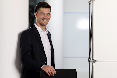 Ein junger Mann in schwarzem Anzug und weißem Hemd steht lächelnd an einer Glaswand in einem modernen Bürogebäude. Er hat dunkles, kurzes Haar und posiert professionell für ein Businessfoto.