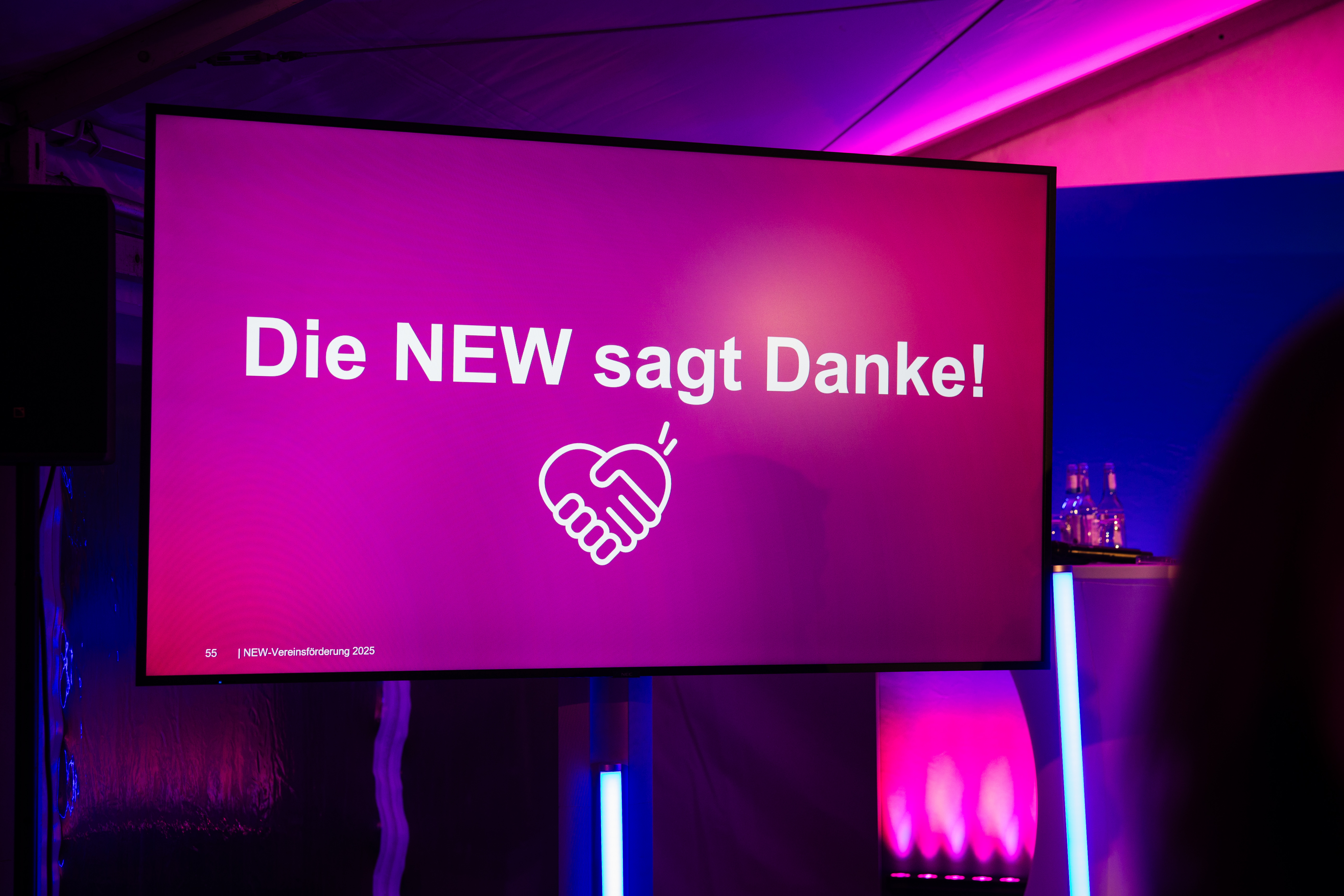  Ein großer Bildschirm mit pinkem Hintergrund zeigt die Aufschrift „Die NEW sagt Danke!“ sowie ein Handschlag-Symbol. Die Szene ist in violettem Licht beleuchtet und wirkt wie eine Veranstaltung oder Präsentation.