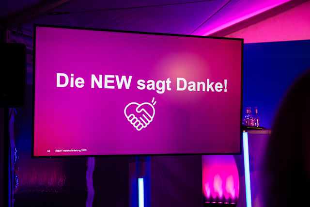 Menschen verbinden, Gemeinschaft stärken. Ein großer Bildschirm mit pinkem Hintergrund zeigt die Aufschrift „Die NEW sagt Danke!“ sowie ein Handschlag-Symbol. Die Szene ist in violettem Licht beleuchtet und wirkt wie eine Veranstaltung oder Präsentation.