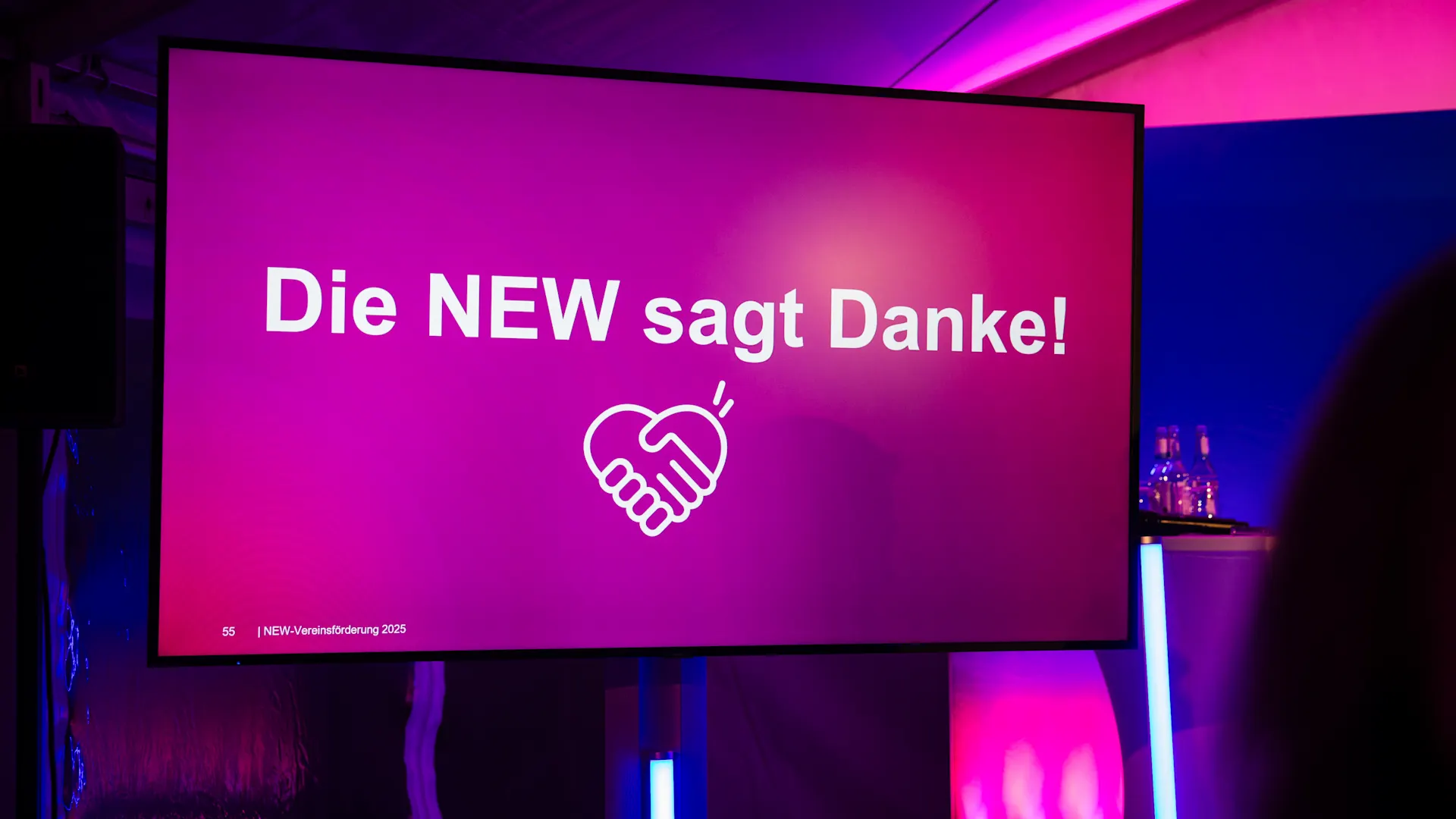 Menschen verbinden, Gemeinschaft stärken. Ein großer Bildschirm mit pinkem Hintergrund zeigt die Aufschrift „Die NEW sagt Danke!“ sowie ein Handschlag-Symbol. Die Szene ist in violettem Licht beleuchtet und wirkt wie eine Veranstaltung oder Präsentation.