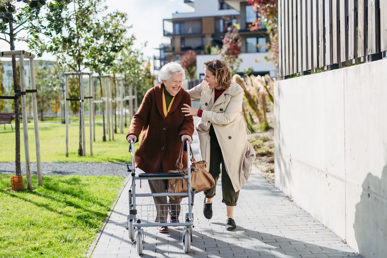 Eine ältere Person nutzt einen Rollator auf einem Gehweg in einer modernen Wohnanlage. Eine weitere Person begleitet die ältere Person und geht neben ihr. Im Hintergrund sind Grünflächen, junge Bäume sowie mehrgeschossige Wohngebäude zu sehen. Die Szene zeigt selbstständige Mobilität und Unterstützung im Alltag in einem barrierearmen Wohnumfeld.