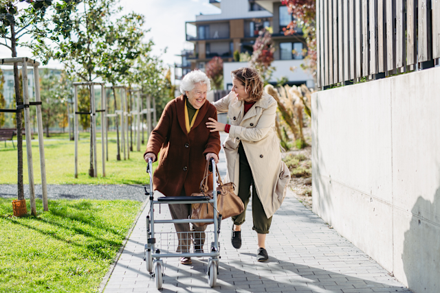 Rollatortag der NEW: Sicher und selbstbewusst unterwegs Eine ältere Person nutzt einen Rollator auf einem Gehweg in einer modernen Wohnanlage. Eine weitere Person begleitet die ältere Person und geht neben ihr. Im Hintergrund sind Grünflächen, junge Bäume sowie mehrgeschossige Wohngebäude zu sehen. Die Szene zeigt selbstständige Mobilität und Unterstützung im Alltag in einem barrierearmen Wohnumfeld.