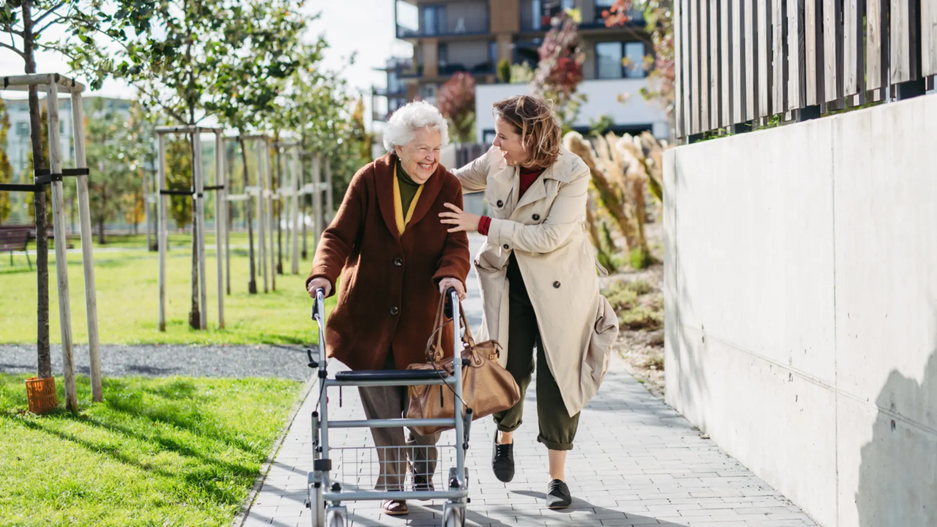 Rollatortag der NEW: Sicher und selbstbewusst unterwegs Eine ältere Person nutzt einen Rollator auf einem Gehweg in einer modernen Wohnanlage. Eine weitere Person begleitet die ältere Person und geht neben ihr. Im Hintergrund sind Grünflächen, junge Bäume sowie mehrgeschossige Wohngebäude zu sehen. Die Szene zeigt selbstständige Mobilität und Unterstützung im Alltag in einem barrierearmen Wohnumfeld.