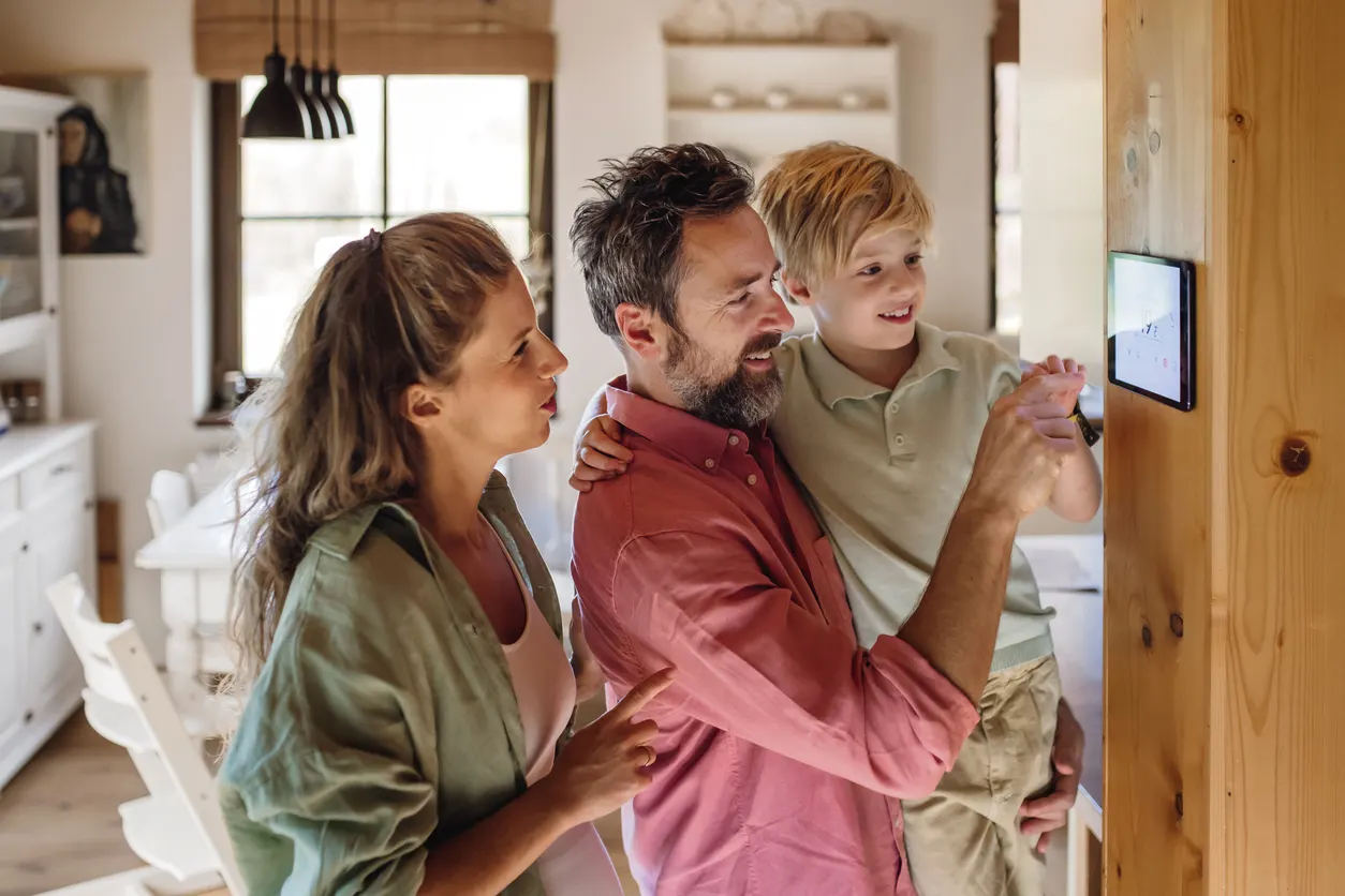 Smart Home Eine junge Familie steht vor einem Smart-Home-Panel an der Wand. Der Vater hält seinen Sohn auf dem Arm, während der Junge auf das Bedienfeld tippt. Die Mutter schaut auf das Display.