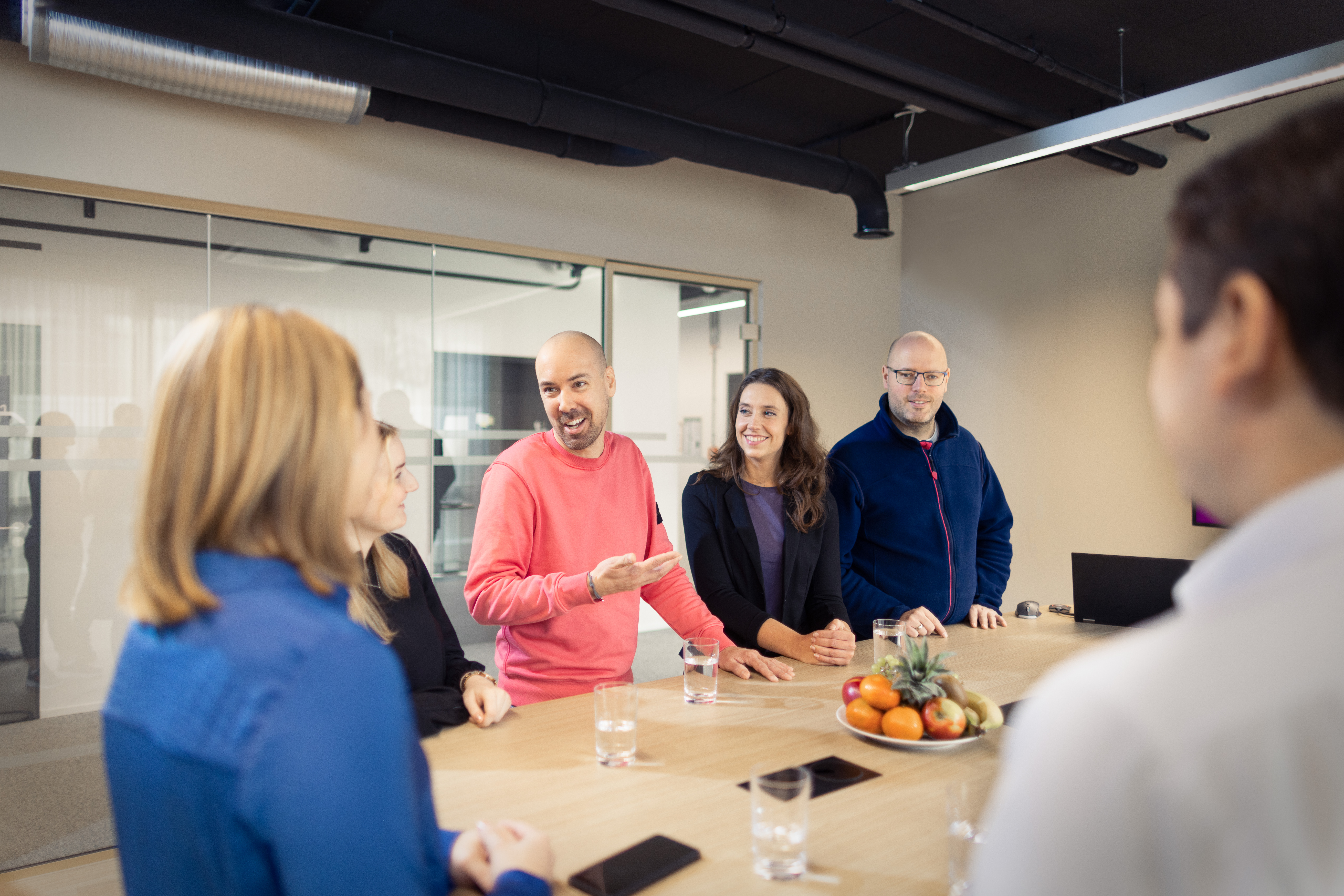 Mehrere Personen stehen in einem Besprechungsraum um einen Konferenztisch und tauschen sich aus. Auf dem Tisch liegen Obst, Wassergläser und Smartphones, während eine Person in einem pinken Pullover spricht und gestikuliert.