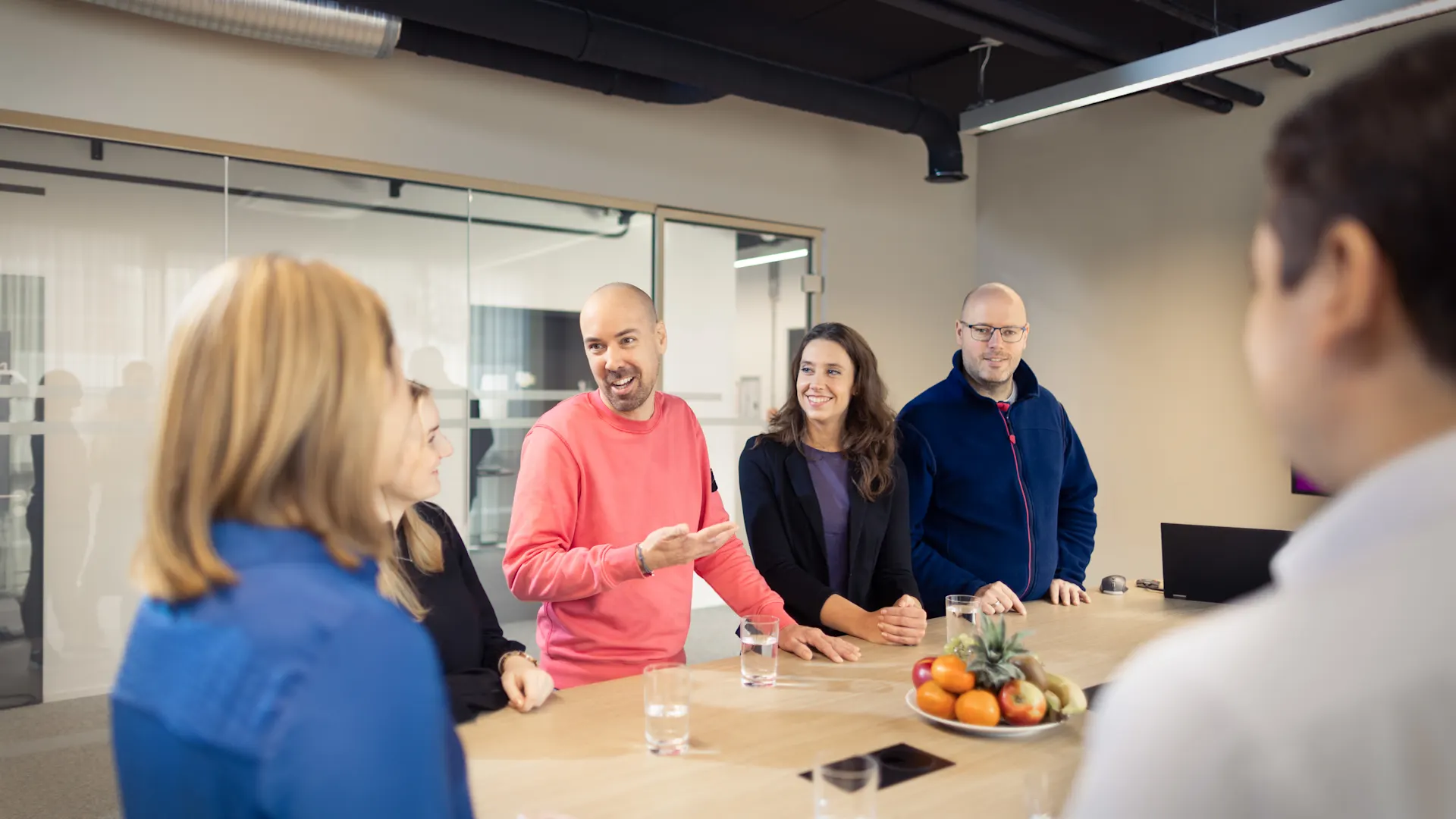 Menschen verbinden, Gemeinschaft stärken. Mehrere Personen stehen in einem Besprechungsraum um einen Konferenztisch und tauschen sich aus. Auf dem Tisch liegen Obst, Wassergläser und Smartphones, während eine Person in einem pinken Pullover spricht und gestikuliert.