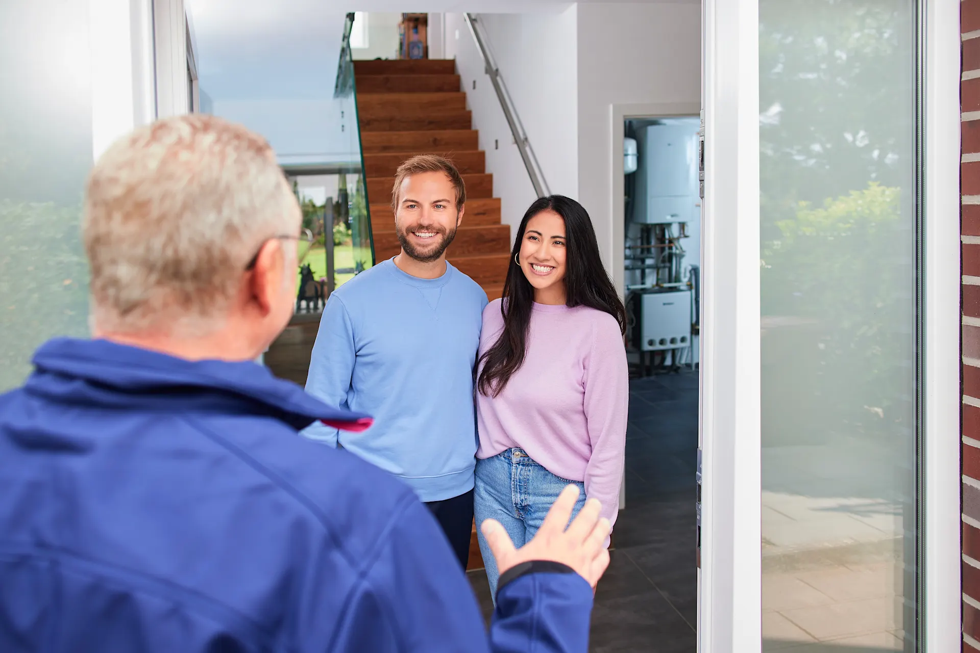 Hausbesuch der NEW Netz Ein Handwerker begrüßt eine junges Paar freundlich in ihrem Eigenheim. Im Hintergrund ist eine geöffnete Tür mit einer modernen Heizungsanlage zu sehen.