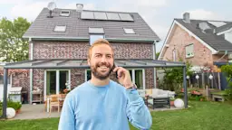 Mann telefoniert vor seinem Einfamilienhaus Zu sehen ist ein Einfamilienhaus, das aus der Perspektive des Gartens fotografiert ist. Der Mann steht im Vordergrund und telefoniert mit einem Smartphone.