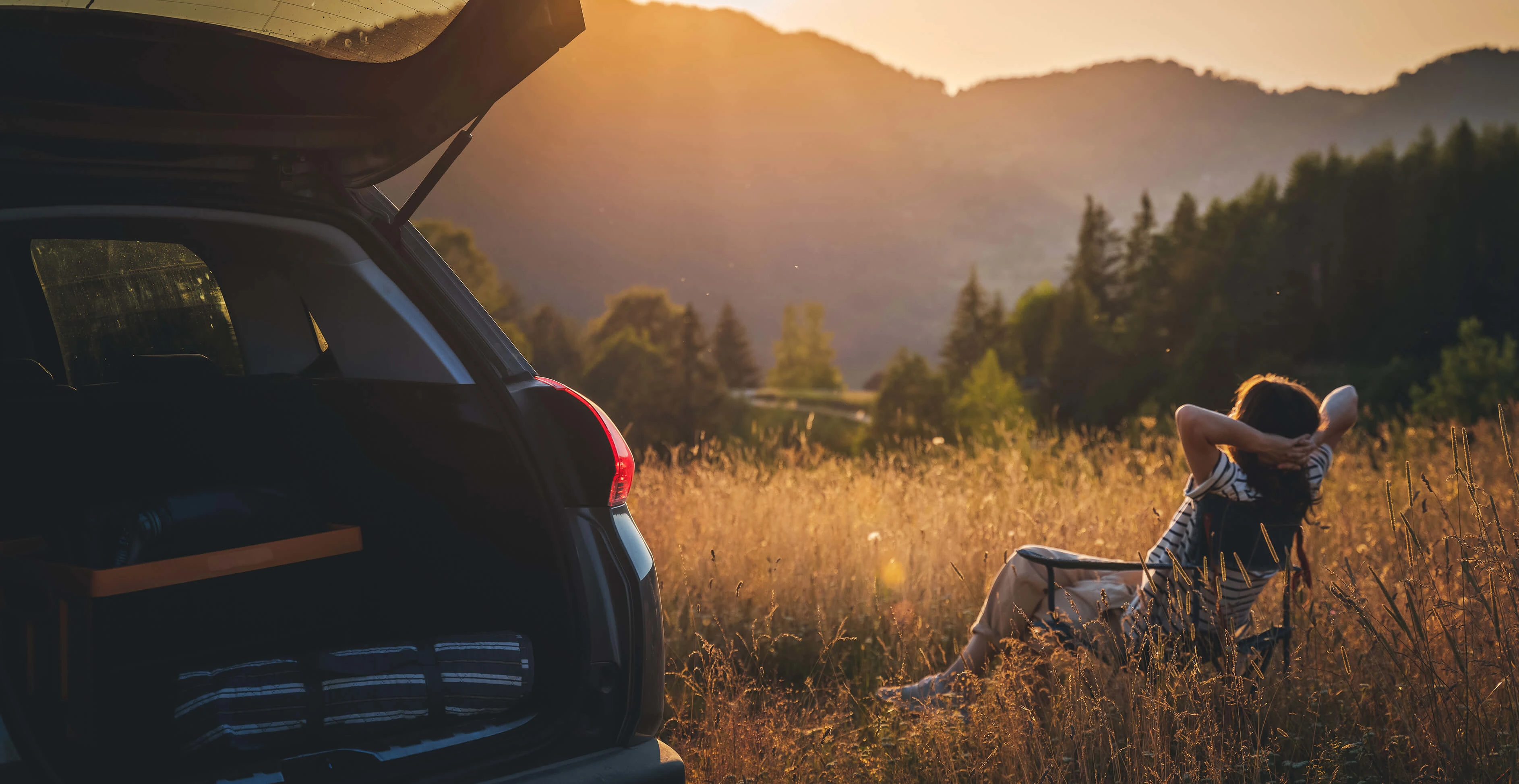 Frau genießt auf ihrem Campingstuhl neben ihrem Auto den Sonnenuntergang