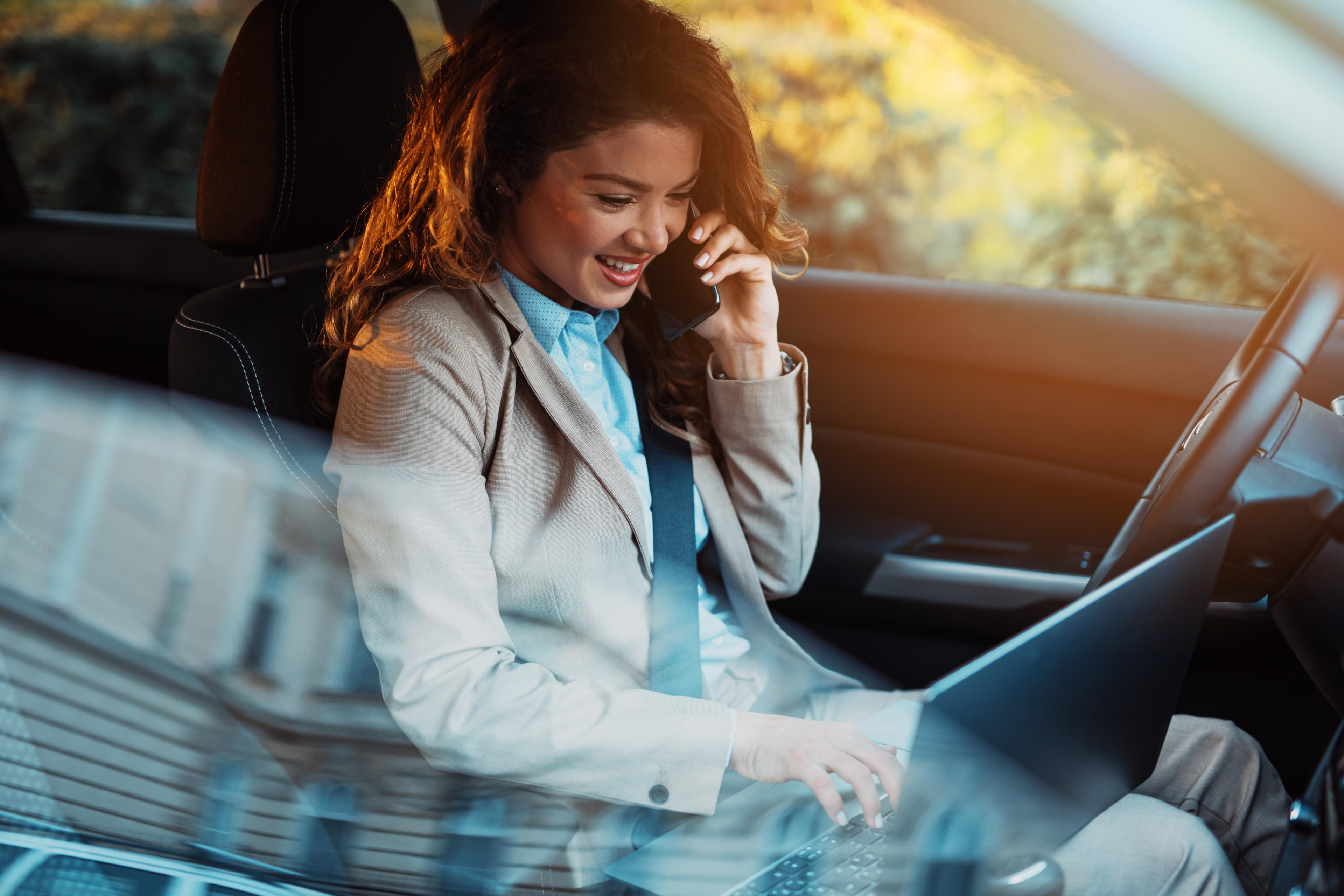 Frau sitzt im Auto und arbeitet mit Smartphone in der Hand und Laptop