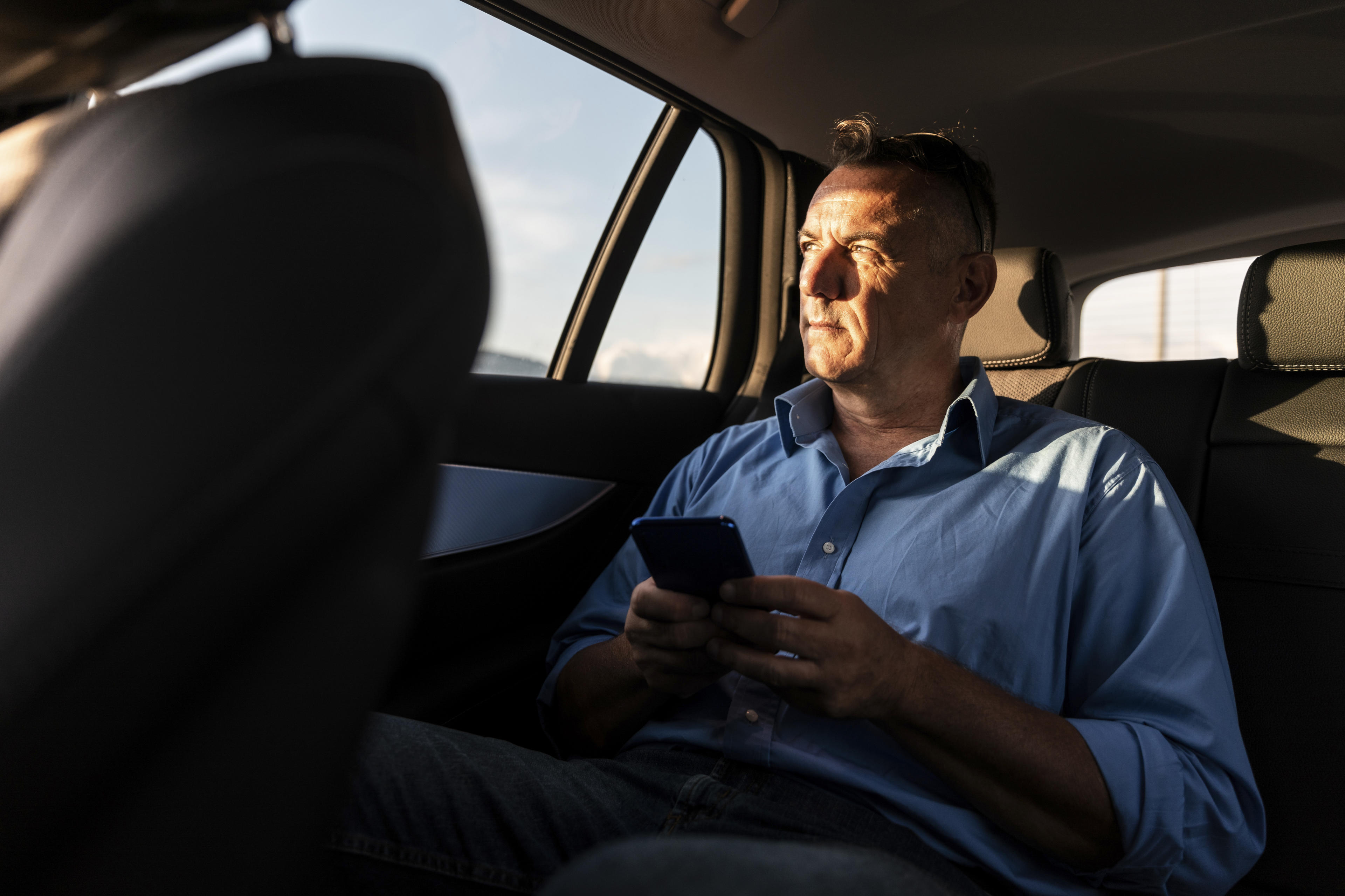 Mann hält Smartphone in der Hand und sitzt am geöffneten Kofferraum seines Autos