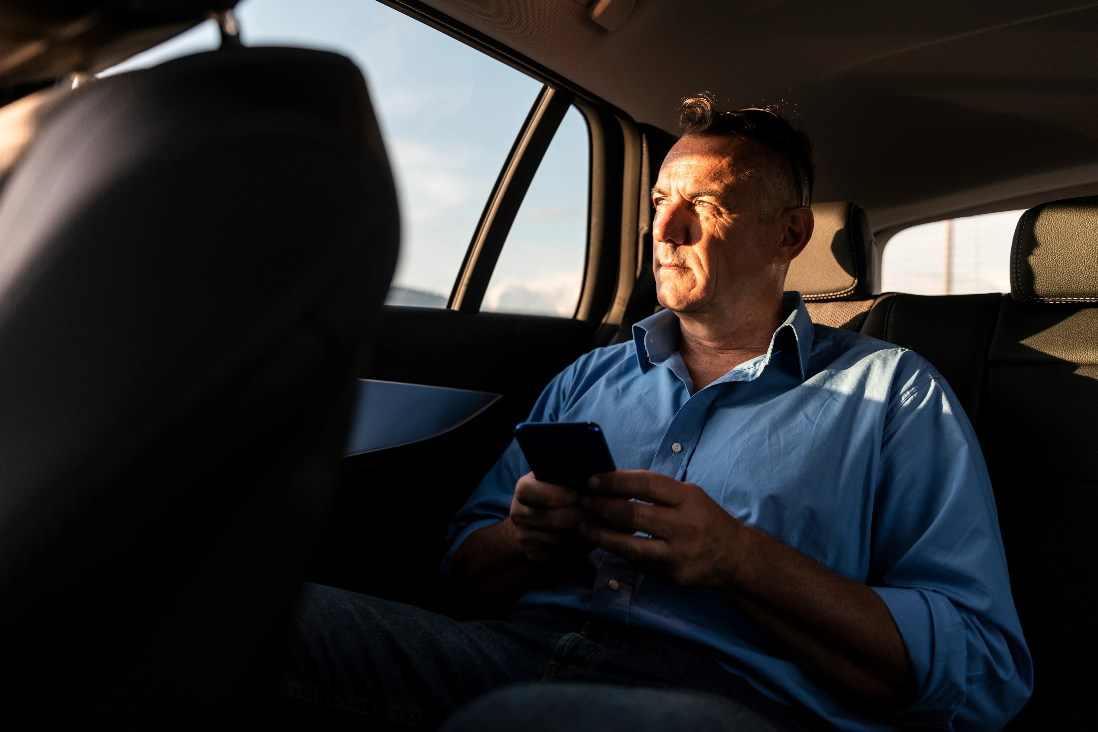 Mann hält Smartphone in der Hand und sitzt am geöffneten Kofferraum seines Autos