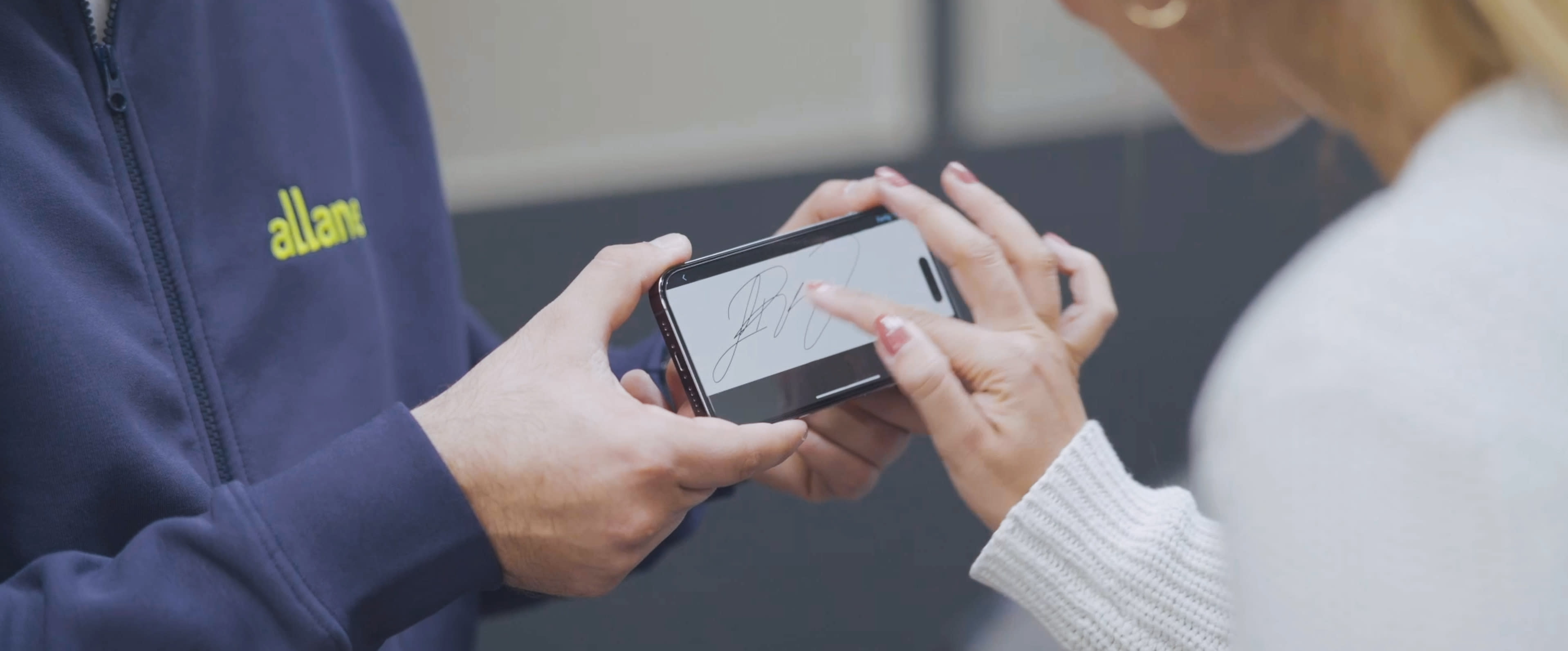 Ein Allane Mitarbeiter hält einer Frau ein Smartphone, auf dem sie mit dem Finger unterschreibt.