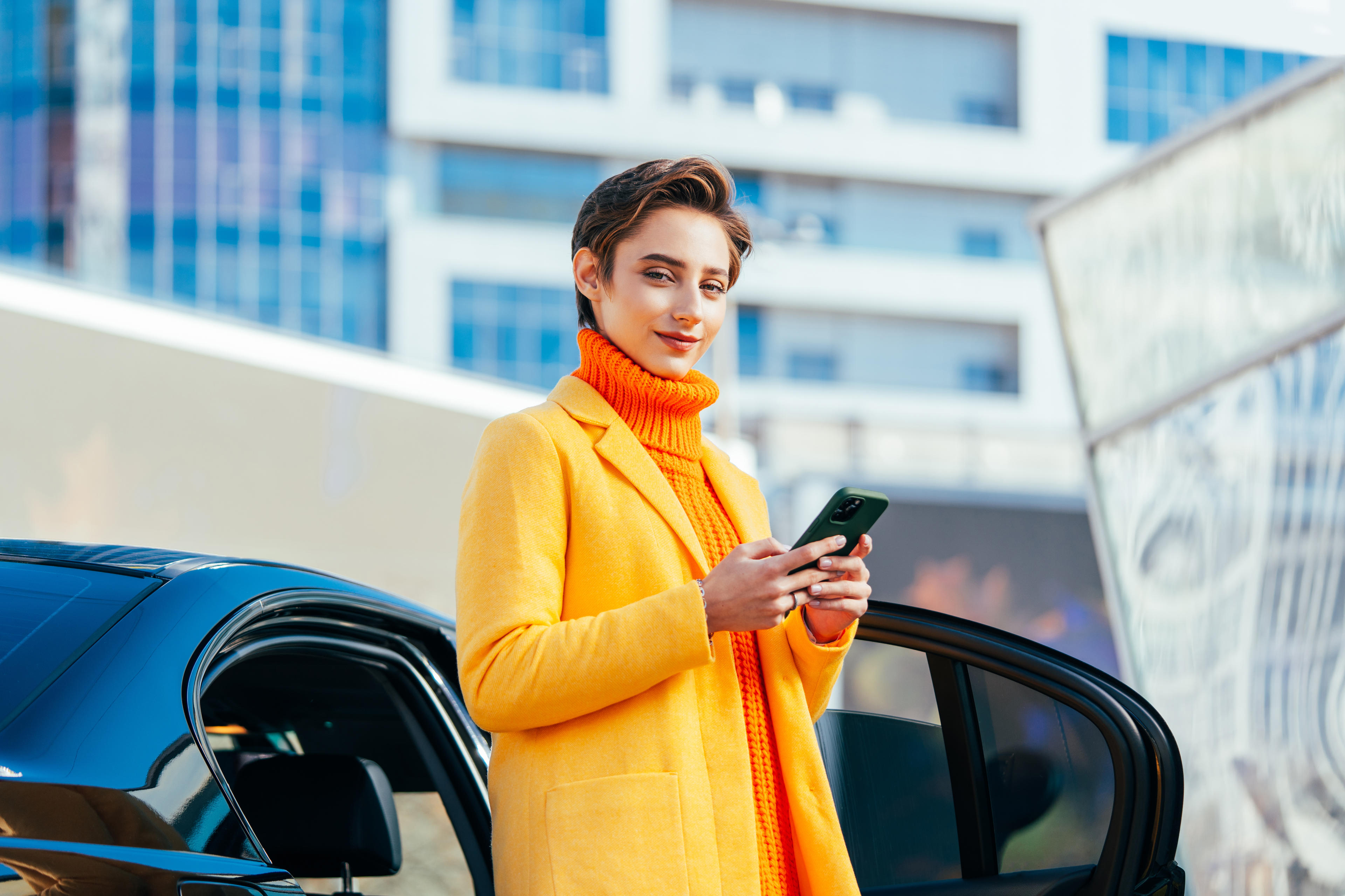 Frau in einem orangen Mantel steht vor einem Auto mit Smarthpone in der Hand