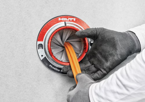 Nahaufnahme einer Hilti Brandschutz-Kabeldurchführung in einer Wand. Behandschuhte Hände führen orange Kabel durch ein kreisförmiges Brandschutzsystem mit flexiblen Dichtlamellen zur Abschottung von Brand- und Rauchabschnitten.