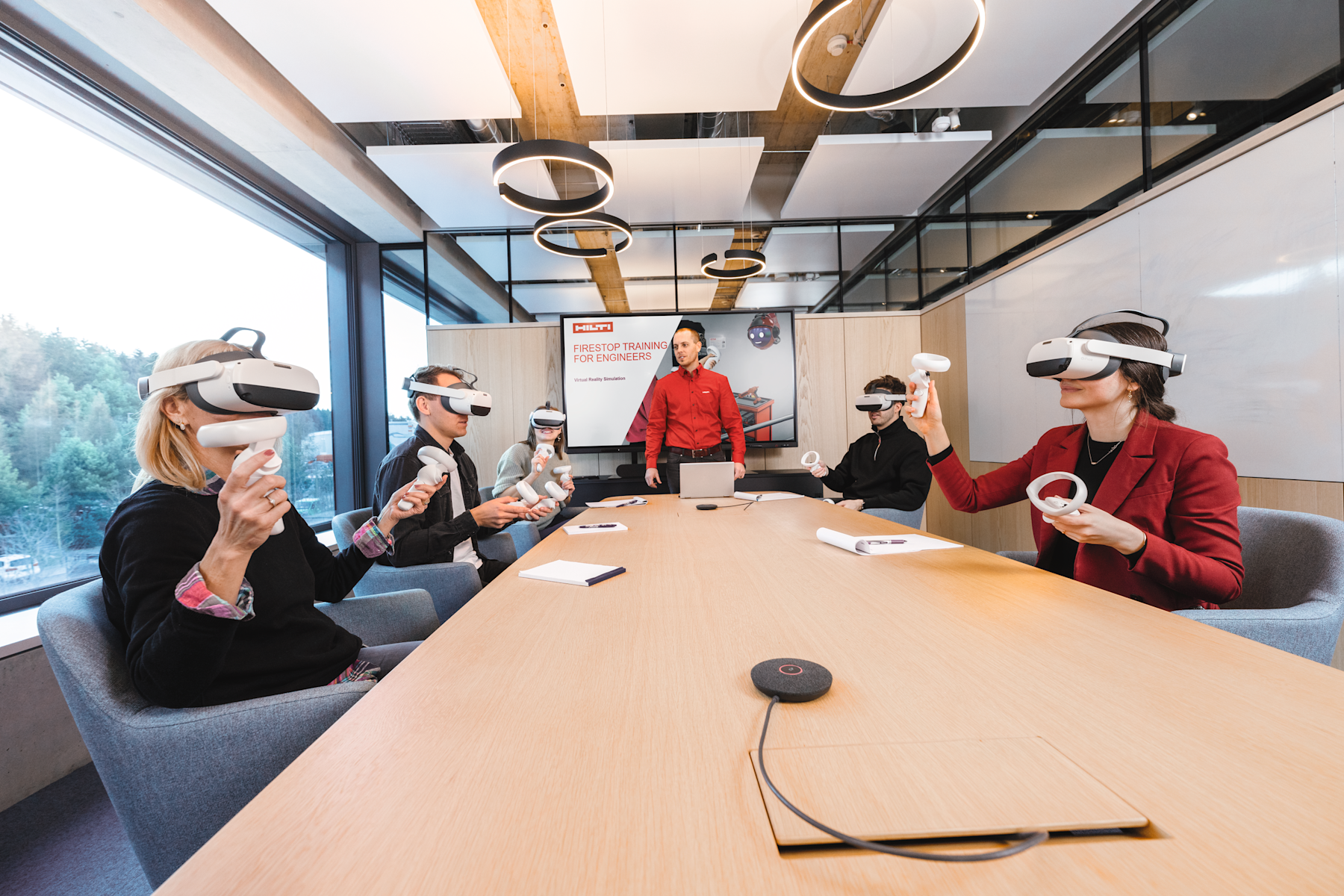 Mehrere Teilnehmende nutzen während einer Schulung Virtual-Reality-Headsets und Handcontroller an einem Besprechungstisch. Vorne im Raum sind eine Präsentation und eine präsentierende Person zu sehen.