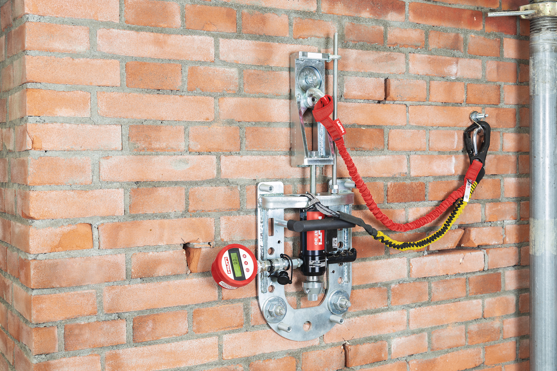 Auszugprüfgerät an einer Ziegelwand mit hydraulischer Belastungseinrichtung, digitaler Hilti-Anzeige und Sicherungsbändern zur Aufbringung und Messung einer Zuglast auf eine Dübelbefestigung.