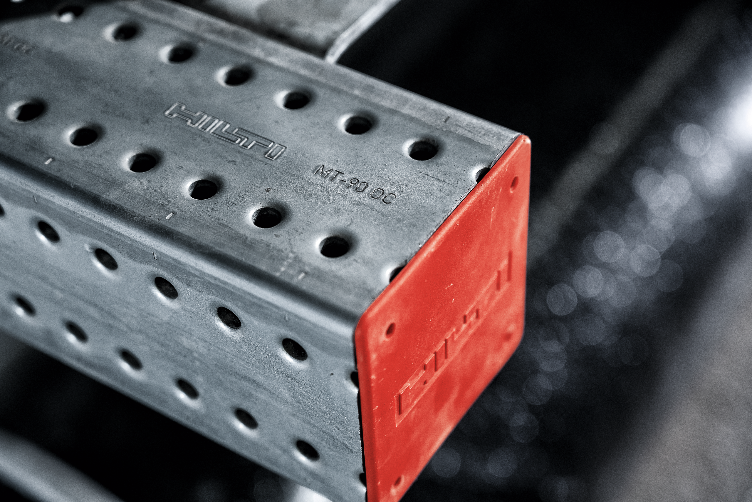 Nahaufnahme einer perforierten Hilti MT-90 OC Montageschiene aus verzinktem Stahl. Das offene Schienenende ist mit einer roten Schutzkappe versehen, während Hilti-Logo und Produktbezeichnung in die Metalloberfläche eingeprägt sind.