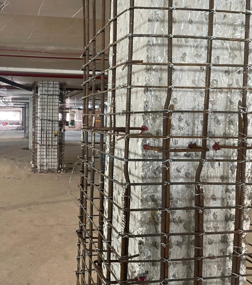 Concrete columns wrapped with steel reinforcing cages for structural strengthening inside a building under construction. Multiple strengthened columns are visible in a large open interior space.