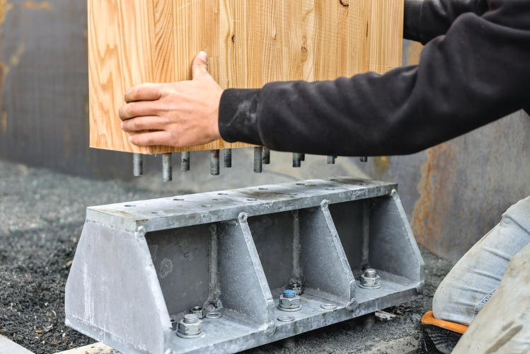 Ein Holzbauteil mit integrierten Gewindestangen wird über einer verzinkten Stahlkonsole auf einem Betonfundament positioniert und zeigt die Montage einer tragenden Holz-Stahl-Beton-Verbindung.