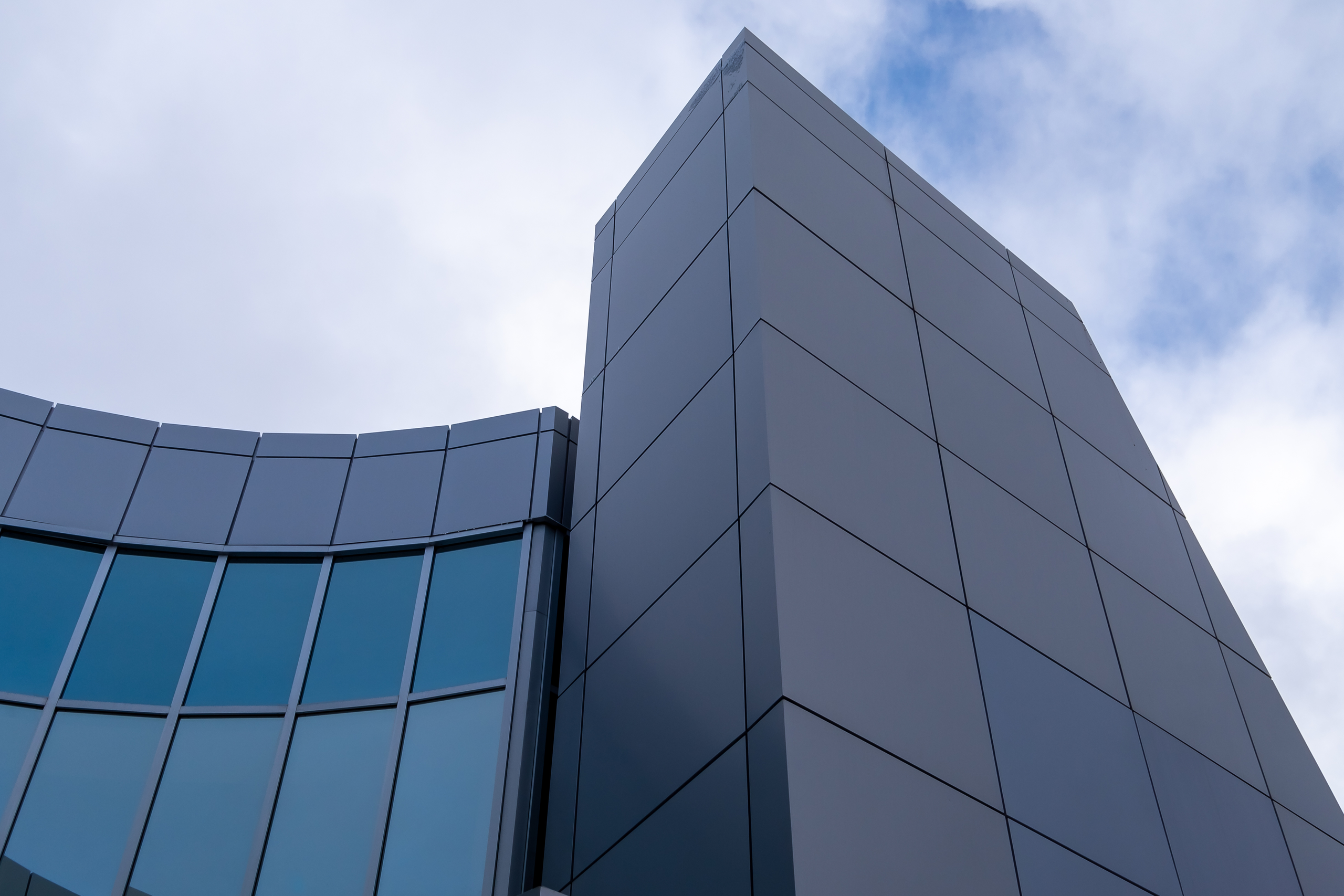 The corner of a grey coloured building made of metal composite panels and green reflective windows. Part of the building has a high tower and a lower curved section made of green windows.