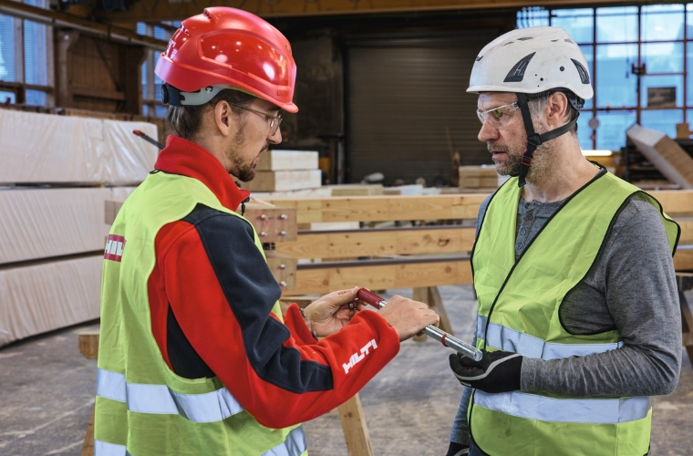 Zwei Fachkräfte prüfen in einer Holzbauwerkstatt eine Stahlstange für eingeklebte Gewindestangen-Verbindungen vor der Montage an vorbereiteten Holzbauteilen.