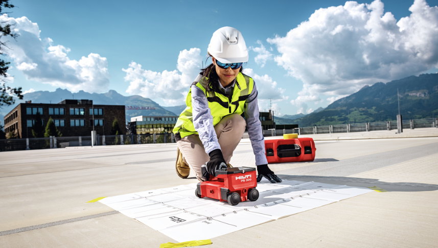 Person på en betongyta använder en röd Hilti PS 300-betongscanner ovanpå tekniska ritningar. I bakgrunden syns byggnader, berg och en molnig himmel. Bilden visar betongscanning inför borr- eller infästningsarbete.
