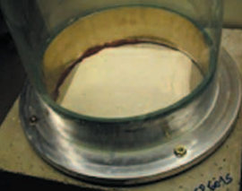 Clear cylindrical chamber mounted on a concrete test slab, with visible sealing ring at the base and a light-colored material inside, used for controlled testing of surface permeability or leakage.
