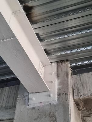Stahlträgeranschluss in einem Bestandsbau. Ein weiß beschichteter Stahlträger ist über eine angeschraubte Stahlplatte auf einer Betonstütze befestigt, unter einer Trapezblechdecke.