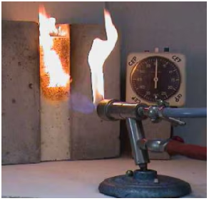 Laboratory fire test with a gas burner flame directed at a wall opening, where flames are visible inside. A thermometer gauge measures heat, showing controlled fire exposure and temperature monitoring.