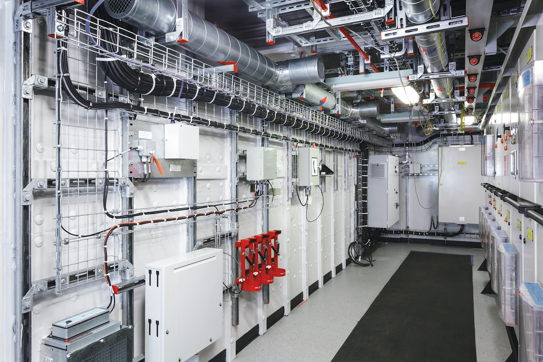 Mechanical room with suspended HVAC ducts, cable trays, and electrical panels, showing organized installations fixed to a concrete ceiling using metal support systems.