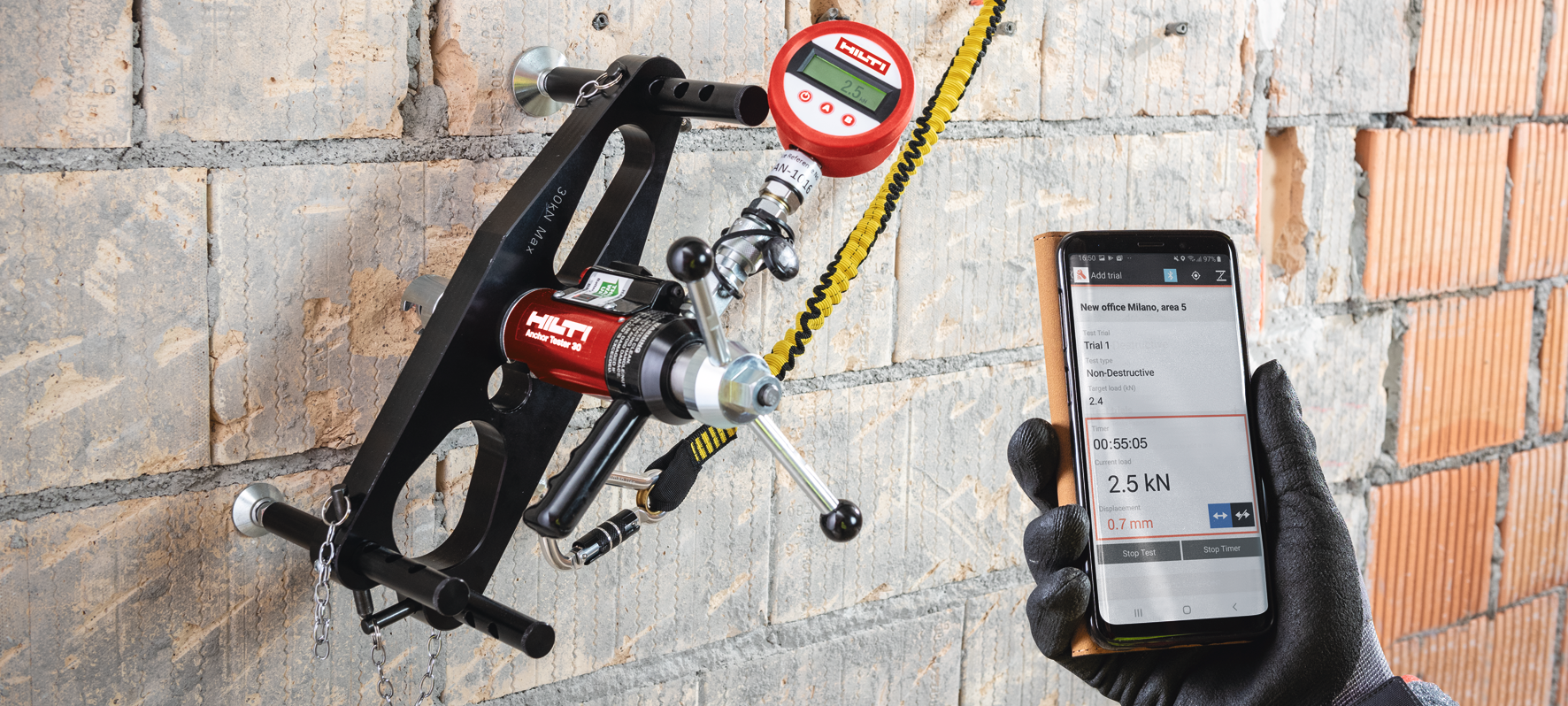 Hilti-Gerät zur Belastungsprüfung eines Dübels an einer Mauerwerkswand. Eine behandschuhte Hand hält ein Smartphone, das einen zerstörungsfreien Test mit 2,5 kN aktueller Last und 0,7 mm Verschiebung anzeigt.