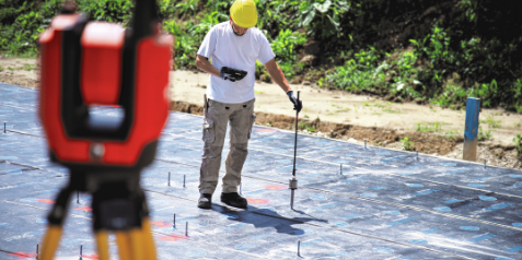 Person wearing a hard hat uses a long installation tool on a concrete slab with anchor bolts visible nearby. A red surveying instrument is blurred in the foreground at an outdoor construction site.