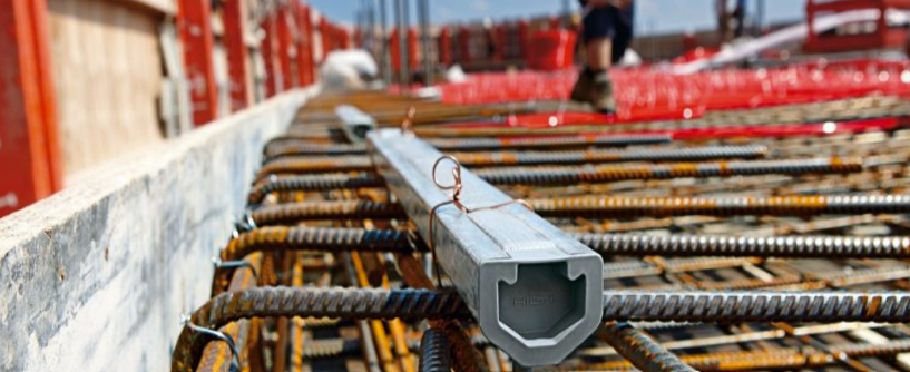 Close-up of steel reinforcement bars arranged in a grid at a construction site, with a hollow rectangular conduit tied on top; blurred worker and red safety barriers visible in the background.
