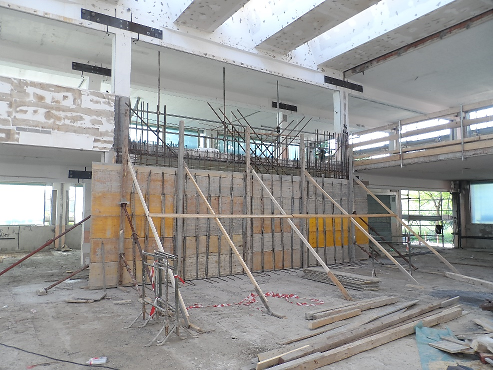 Concrete shear wall under construction inside a building, with vertical rebar protruding at the top, temporary wooden formwork, and diagonal braces supporting the structure.