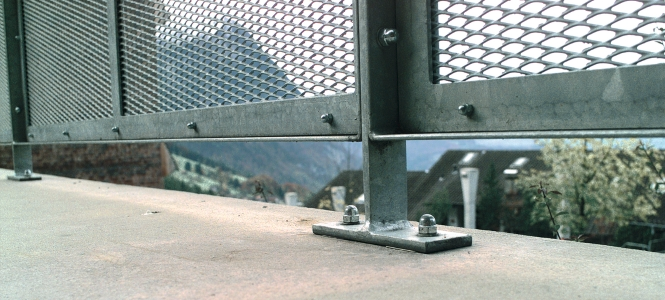 Metallgeländer mit Streckmetallfüllung, befestigt über eine angeschweißte Fußplatte und mechanische Verankerungen auf einer Betonplatte.