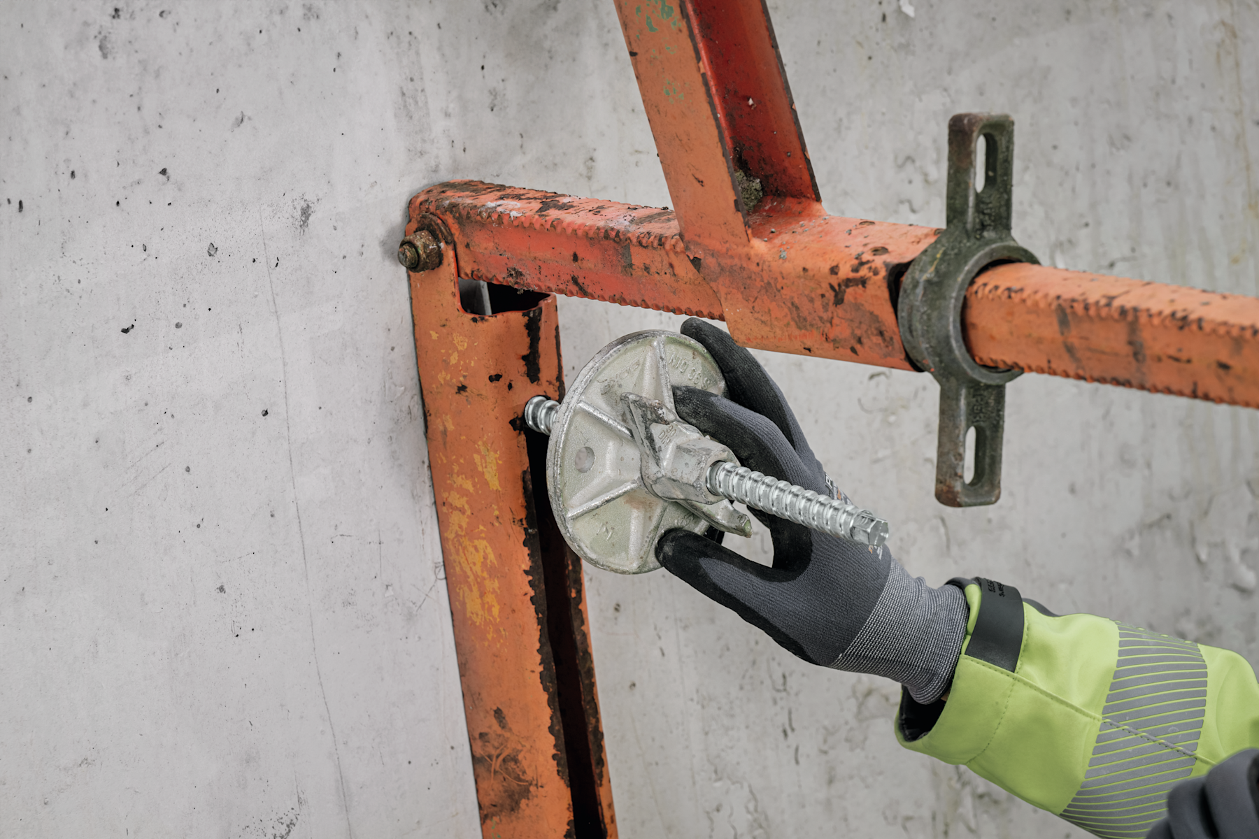 Close-up of a threaded anchor rod with a circular plate being installed by a gloved hand into an orange steel support bracket fixed against a concrete wall.