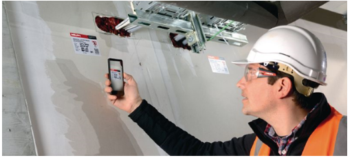 Construction professional using a smartphone to inspect and document firestop penetrations around cable trays passing through a fire-rated wall.