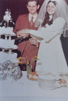 An old, physical photograph of Gwen and John at their wedding. Gwen is wearing a long-sleeved wedding dress and veil, and Tom is wearing a brown suit. They are cutting their wedding cake.