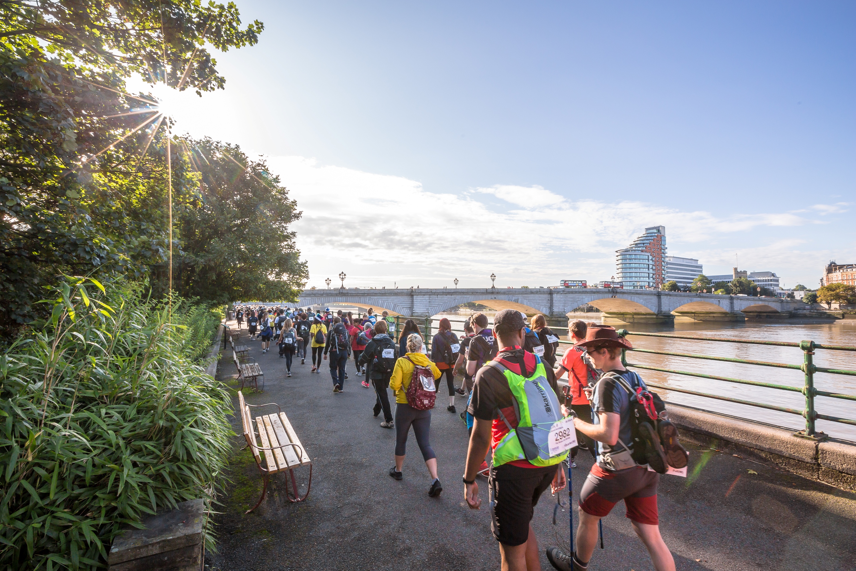 A large group of people walking along the river path with London's Putney bridge in the background.