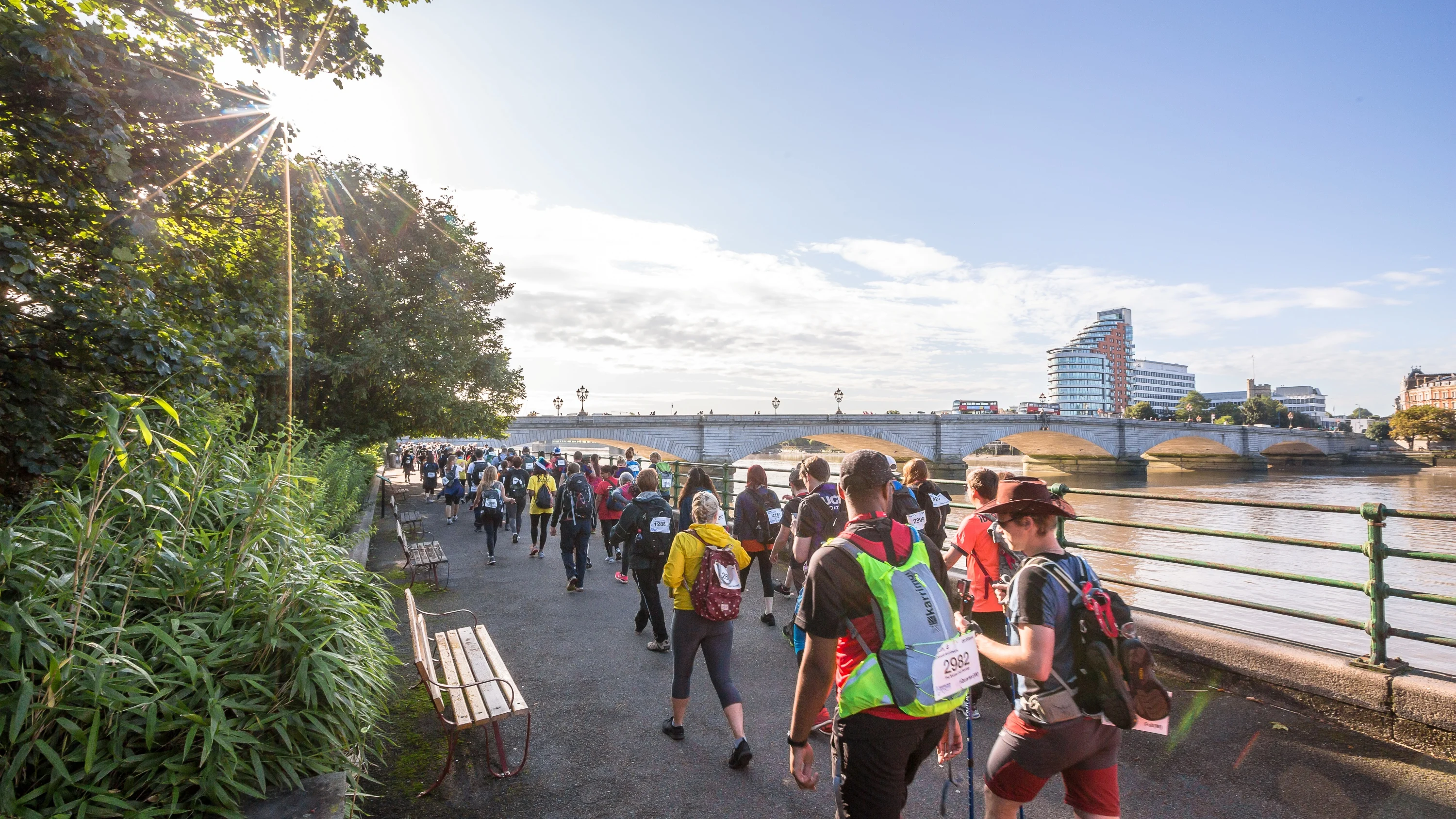 A large group of people walking along the river path with London's Putney bridge in the background.