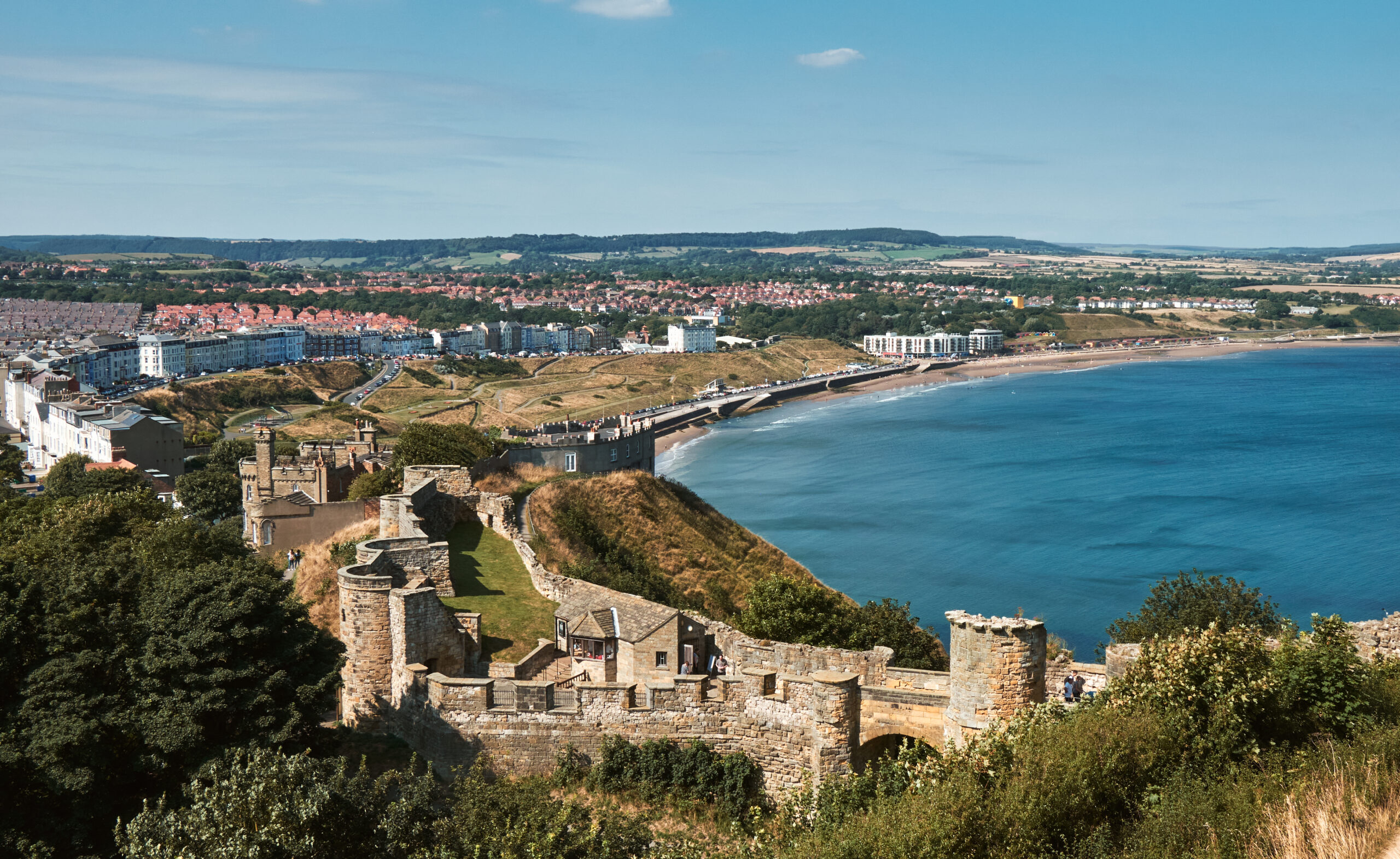 An aerial view of Scarborough on a sunny day with views of the beach, Scarborough castle and distant hills.