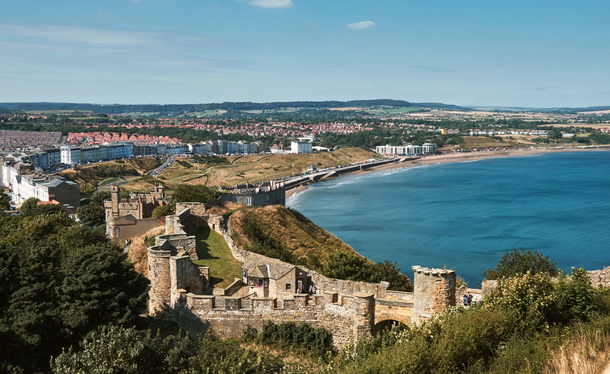 An aerial view of Scarborough on a sunny day with views of the beach, Scarborough castle and distant hills.