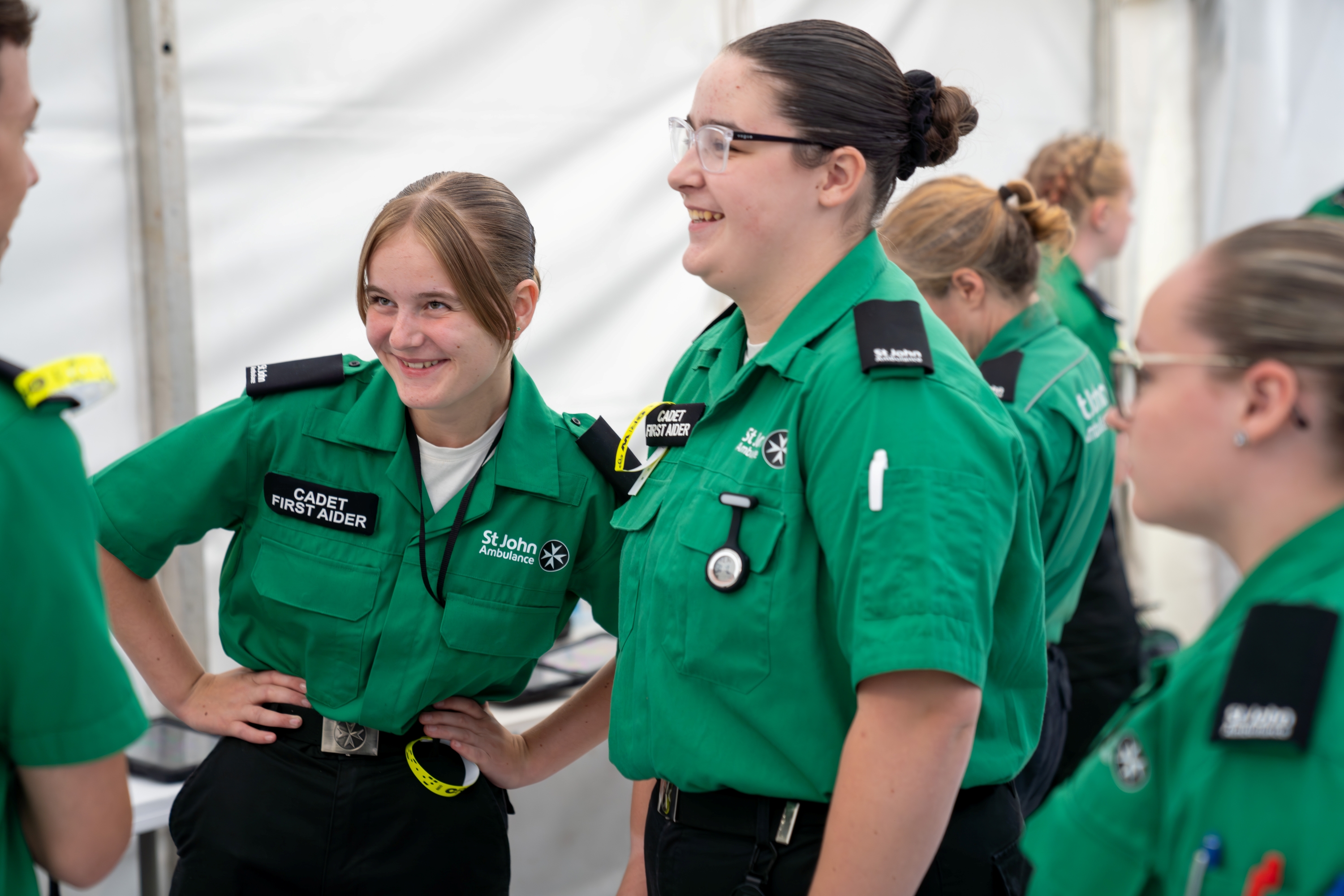 A group of Cadet First Aiders standing around in a white tent. Two are smiling with their hands on their hips in front of another volunteer. 