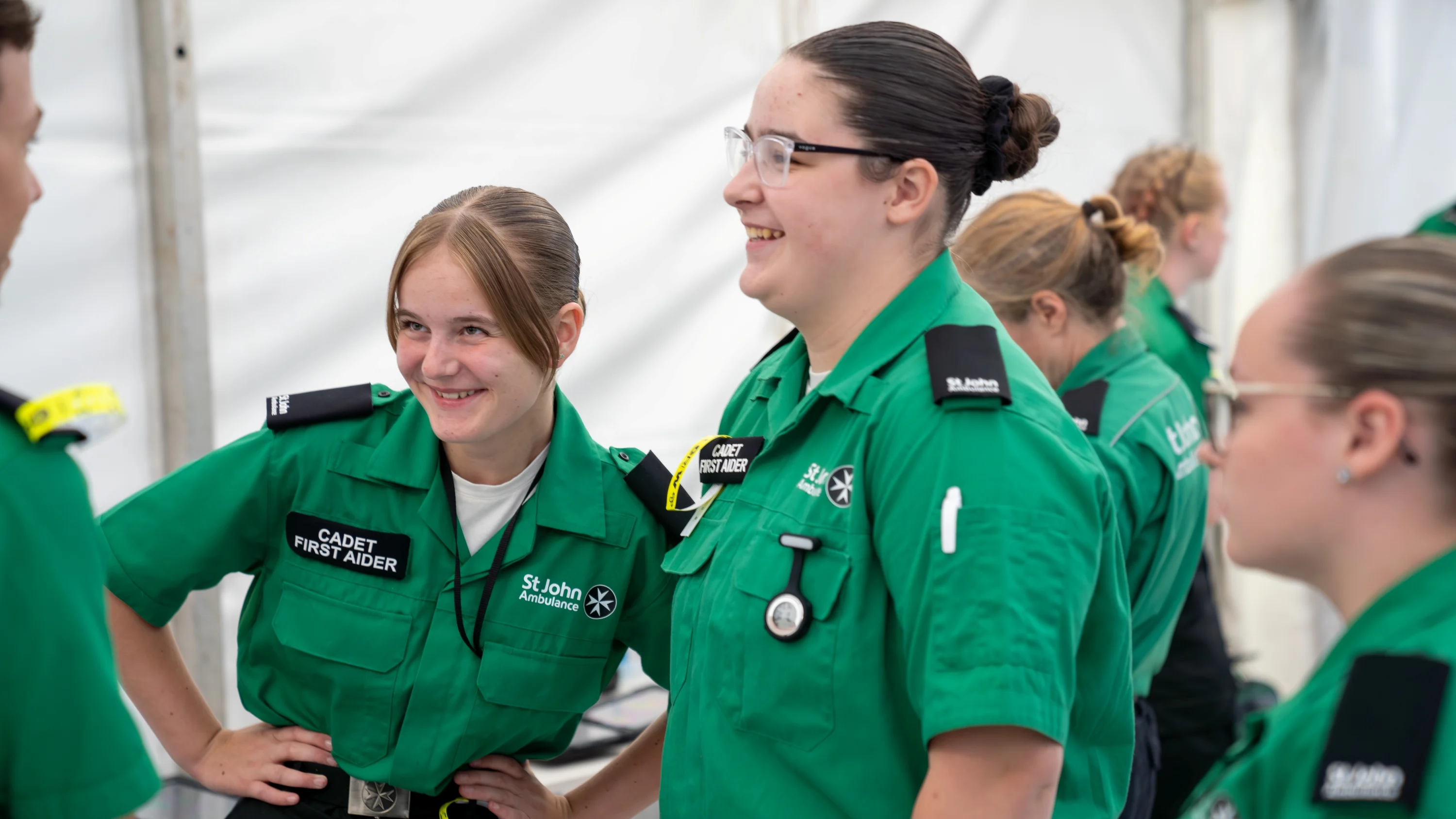 A group of Cadet First Aiders standing around in a white tent. Two are smiling with their hands on their hips in front of another volunteer.